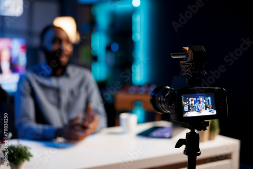 Focus on professional camera capturing footage of BIPOC man chatting with online audience. Viral content creator in blurry background talking with fans, filming with high quality recording tech
