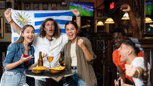Happy international sport supporters holding up the flag of Uruguay and drinking beer with snacks in the pub