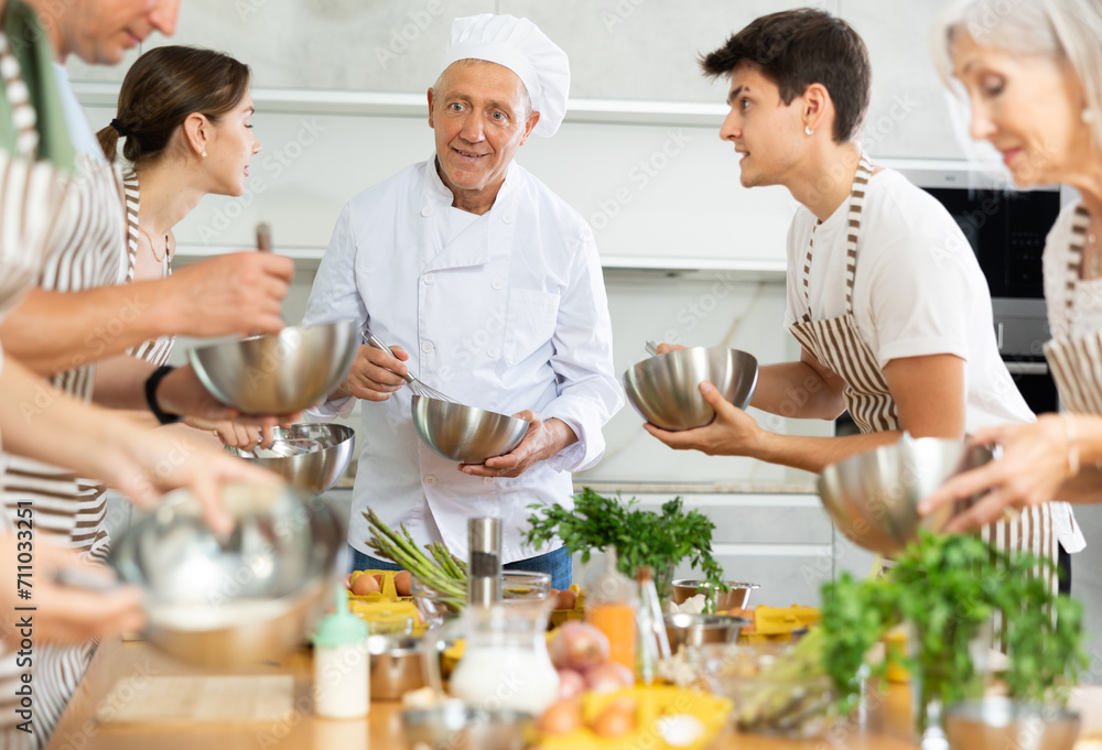 Positive experienced senior chef giving culinary classes to group of interested men and women of different ages, teaching to cook deliciously..