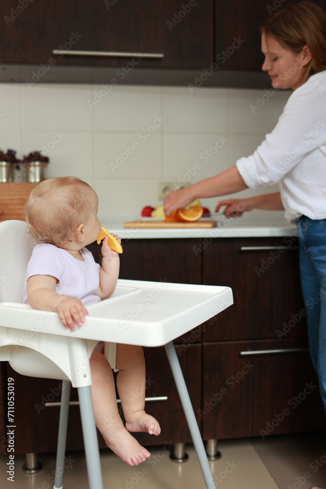 Adorable baby girl eating orange. Little daughter sitting in feeding chair and eating slice of orange at kitchen and playing with mom. Juicy fruits oranges are for a healthy diet. Happy childhood. Adorable baby girl eating orange. Little daughter sitting in feeding chair and eating slice of orange at kitchen and playing with mom. Juicy fruits oranges are for a healthy diet. Happy childhood.