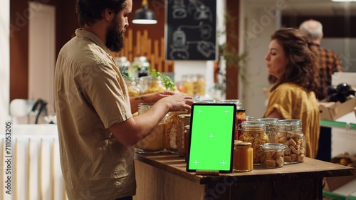 Client shops next to chroma key tablet with copy space used as commercial sign in zero waste supermarket. Promotional ad on green screen device in food store with products in nonpolluting package photo