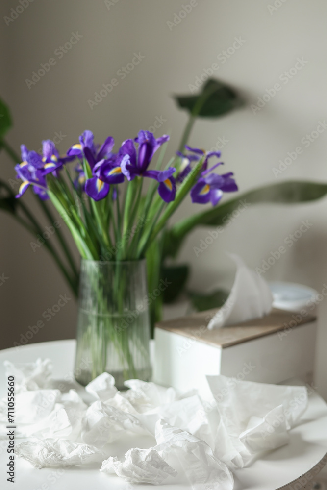 Obraz premium Concept of seasonal allergy to flower. Bouquet of purple iris flowers in a glass vase on table and many used napkins at home interior. Allergic rhinitis symptom of odor pollen flowering