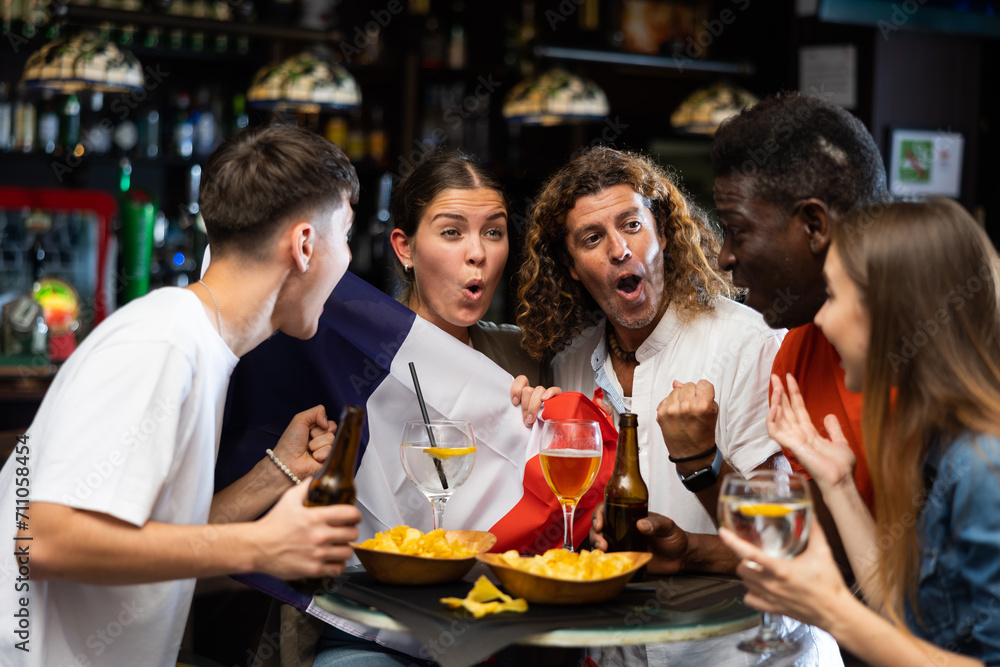 Obraz premium Group of cheerful young adult women and men having fun in sports bar, watching game on TV and rooting for favorite French team