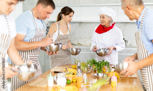 Pleased mature woman cook conducting culinary classes for learners standing around kitchen table with whisk and bowl in hands