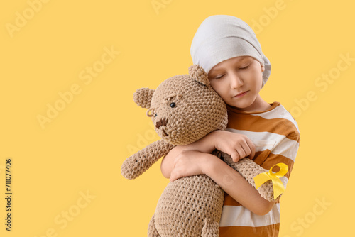 Cute little boy after chemotherapy with yellow ribbon and teddy bear on color background. Childhood cancer awareness concept photo