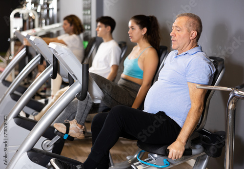 Elderly man, along with other people, is engaged on an exercise bike in the gym