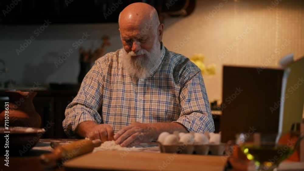 An elderly man with a beard sits at the kitchen table and looks at a recipe book on a stand. The man adds water from a clay jug to the dough, which he kneads with his hands and looks at the book An elderly man with a beard sits at the kitchen table and looks at a recipe book on a stand. The man adds water from a clay jug to the dough, which he kneads with his hands and looks at the book
