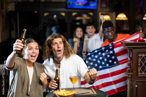 Man and woman, USA football team fans, spending time in bar, drinking bear and screaming chants. People with state flag in pub.