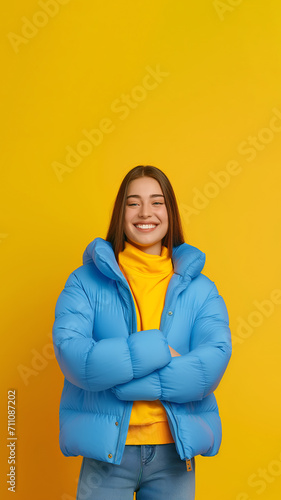 Smiling happy brunette teenage girl wears blue puffer jacket and yellow turtleneck, crossed hands, blue jeans, isolated on yellow backdrop, copy space, vertical 9:16 © Zoran Karapancev
