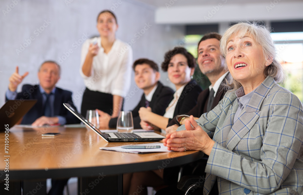 Cheerful elderly female office worker sitting at table with colleagues, watching presentation and laughing during work meeting