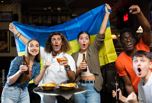 Group of Ukraine football team fans spending time in bar, drinking bear and having fun. People with state flag in pub.