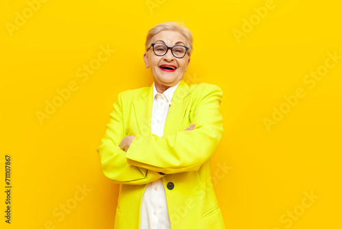 old grandmother in business clothes blazer and glasses laughing crossing her arms on a yellow isolated background, elderly businesswoman in a suit