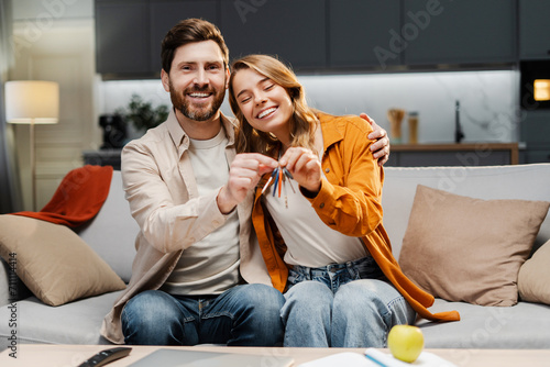 Happy lovely couple holding keys from new home looking at camera sitting on comfortable sofa. Smiling middle aged man and woman buying apartments photo