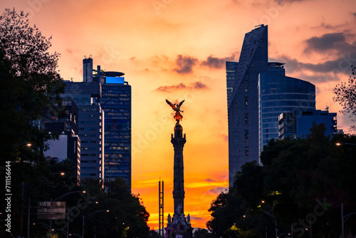 The Angel of Independence statue placed on Promenade of the Reform in Mexico city between tall buildings against colorful orange sunset sky in evening photo