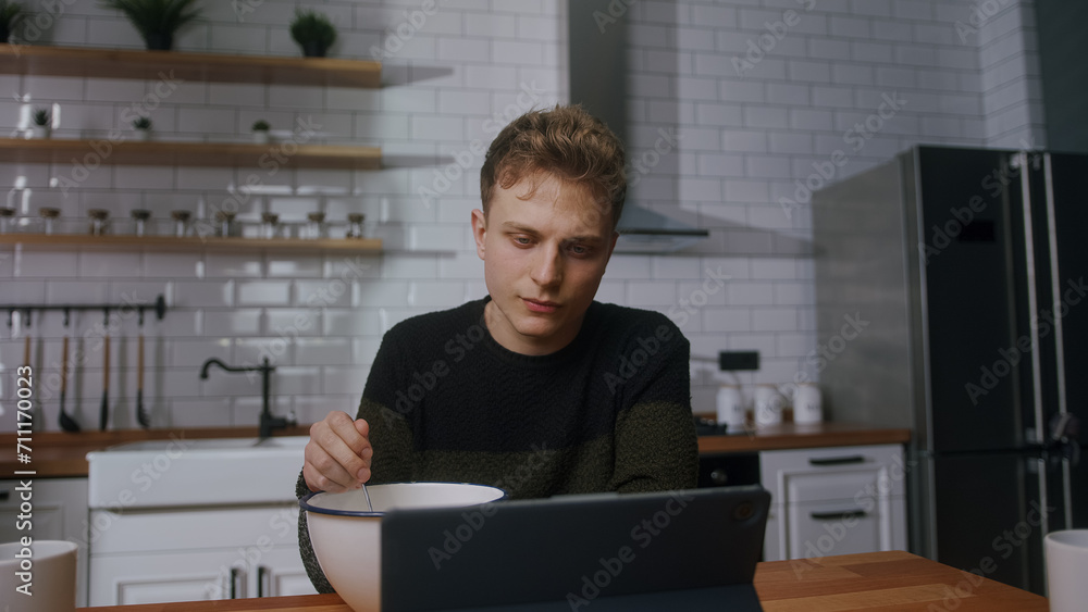 Young man reluctant to eat pushing a bowl full of cereal while sitting in the kitchen watching a video on a tablet computer
Young man reluctant to eat pushing a bowl full of cereal while sitting in the kitchen watching a video on a tablet computer