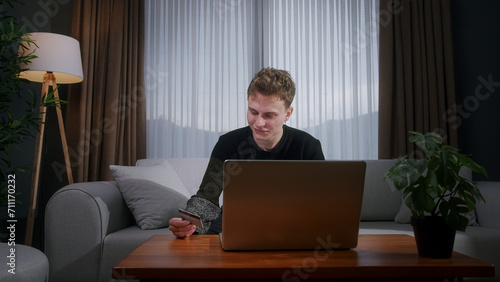 Smiling young man sitting enter credit card number on laptop for makes secure easy distant electronic payment while sitting on sofa at home. Pay for goods services on internet, transactions 