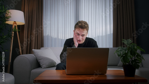 Young male sitting on sofa at home while using laptop having anxiety and stress. Upset young man reacting to loss, bad news on laptop 