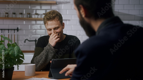 Two young co-worker men are sitting in the kitchen having a fun chat and laughing. Taking a break at work, technology company workers, relaxing time. OTS shot	