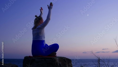 Middle aged woman meditating folding hands in namaste in lotus pose by sea. Blonde female sitting on stone practicing yoga admiring wild sunset nature. Sport recreation mental spirit practice concept. photo