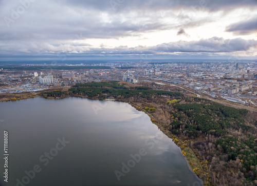 Autumn forest on lake shore at sunset and city on horizon, aerial view