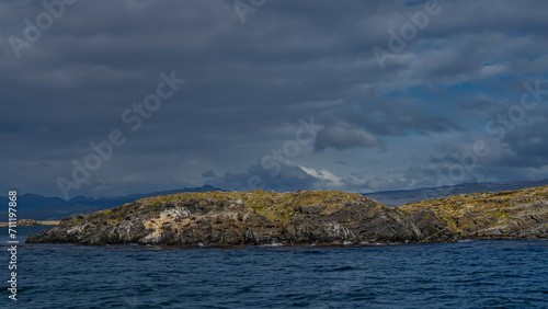 A small rocky island with sparse vegetation in the Beagle Channel. A group of sea lions lies on the cliffs slope. Cormorants fly. Ripples on the blue water. Clouds in the sky. Isla de los lobos.