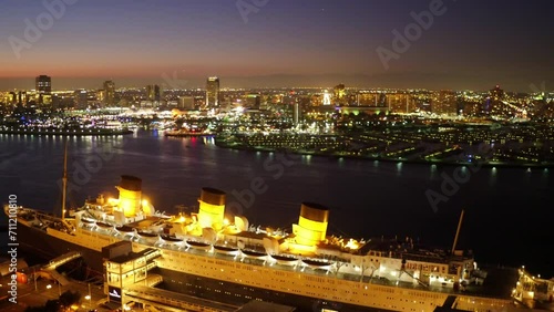 Aerial Panning Shot Of The Queen Mary Cruise Ship Moored In Sea By Illuminated City Against Sky At Night - Long Beach, California