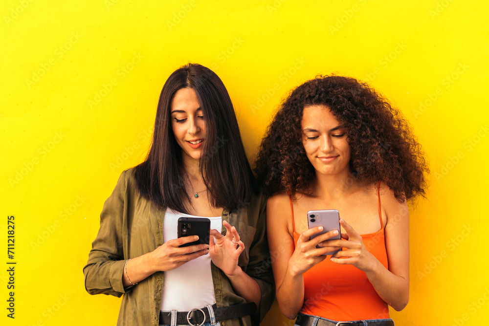 afro and caucasian women watching mobile leaning on a yellow wall afro and caucasian women watching mobile leaning on a yellow wall