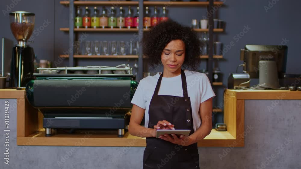 African American female barista holding a tablet in her hand, beautiful young black female barista wearing an apron in a coffee shop, young woman in the middle shot in a coffee shop. African American female barista holding a tablet in her hand, beautiful young black female barista wearing an apron in a coffee shop, young woman in the middle shot in a coffee shop.