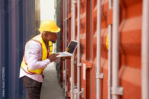 black african man wears hardhat and vest holding laptop computer checking electronic seal on container cargo,young male technician working in shipping yard,concept of container cargo shipping,industry