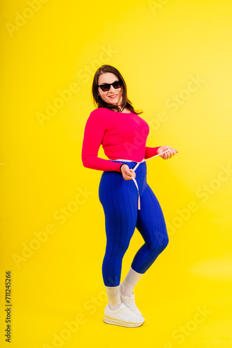 Pretty female with excess weight in sporty top measuring waist over white yellow background