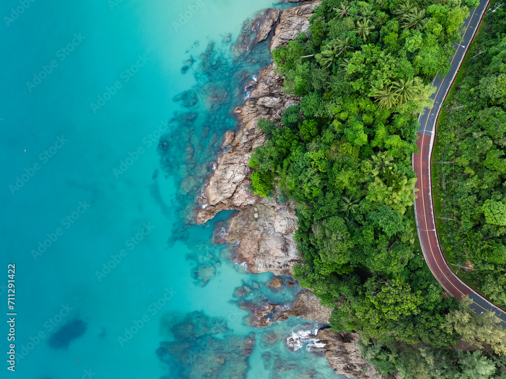 View from above, stunning aerial view of a road that runs alongside a rocky coastline surrounded by palm trees and bathed by a turquoise water. Phuket, Thailand.