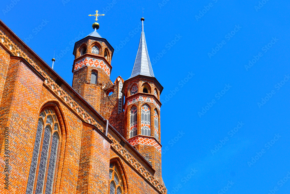 Fototapeta premium Cathedral towers with a cross on top and red brick's wall of catholic church, in the background of blue sky. Sunny summer day. Poland, Torun, August 2023.