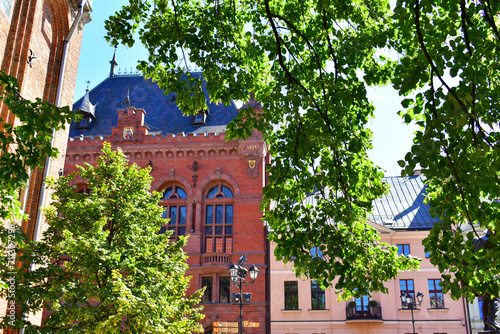 Old buildings made of red brick, decorative elements on the facade, high arched windows and blue roof. Green branches of the trees around. Torun, Poland, August 2023 