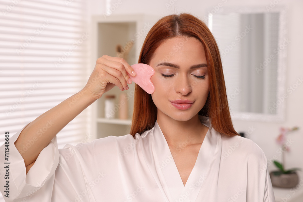 Young woman massaging her face with rose quartz gua sha tool in bathroom Young woman massaging her face with rose quartz gua sha tool in bathroom