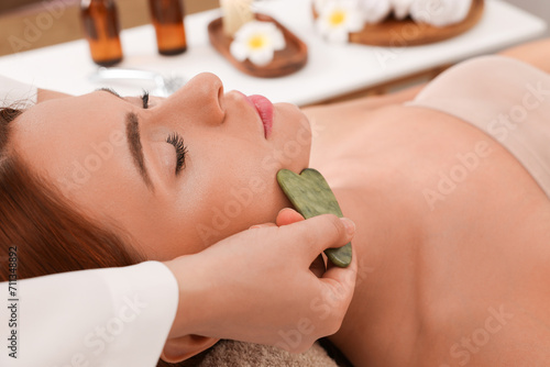 Young woman receiving facial massage with jade gua sha tool in beauty salon, closeup photo