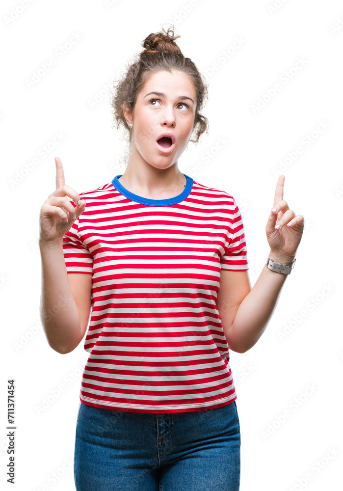 Beautiful brunette curly hair young girl wearing casual look over isolated background amazed and surprised looking up and pointing with fingers and raised arms. Beautiful brunette curly hair young girl wearing casual look over isolated background amazed and surprised looking up and pointing with fingers and raised arms.