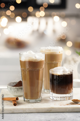 Three tall glasses with warm coffee drink with cinnamon  whipped milk foam and caramel on a marble board with bokeh fairy lights in the background