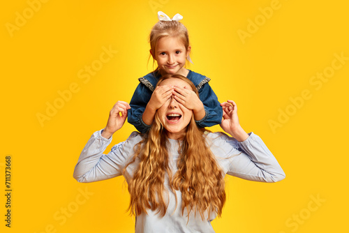 Surprise. Little girl, daughter covering mother's eyes against yellow studio background. Celebrating women's holiday. Concept of Mother's Day, International Happiness Day, family, motherhood photo