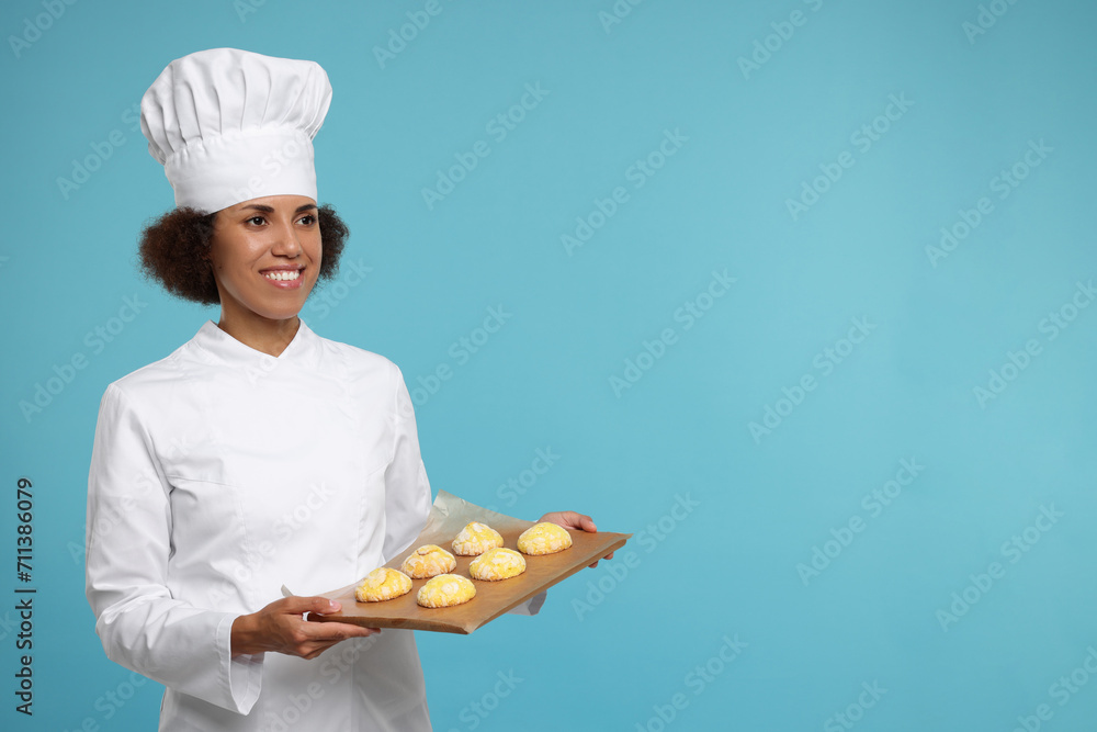 Happy female chef in uniform holding board with cookies on light blue background. Space for text Happy female chef in uniform holding board with cookies on light blue background. Space for text