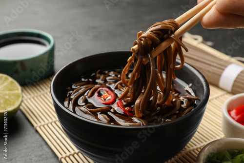 Woman eating delicious buckwheat noodle (soba) soup with chopsticks at grey table, closeup