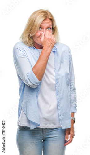 Middle age blonde woman over isolated background smelling something stinky and disgusting, intolerable smell, holding breath with fingers on nose. Bad smells concept.