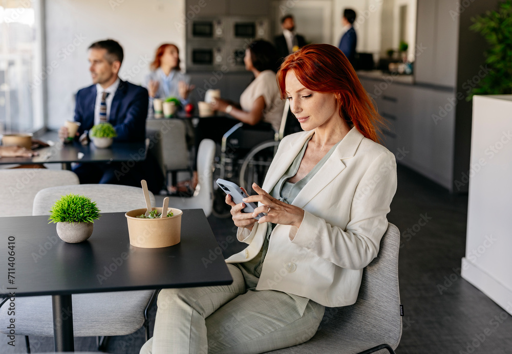 business office, coffee break, pretty red-haired young woman eating on break while looking at her smart phone, colleagues in the background