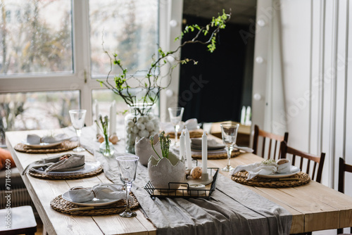 Setting table for Easter dinner with candle, ceramic plates with easter eggs in nest, bunny made of linen napkin. Holidays celebration concept. Decor with vase with twig, branch, leave at home. Set up