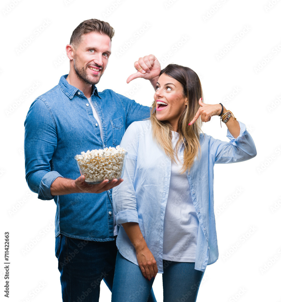 Young couple in love eating popcorn over isolated background with surprise face pointing finger to himself
