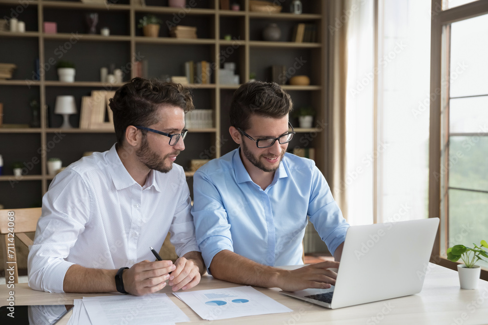 Two male twins sit at desk using laptop, millennial 35s brothers having business partnership, joint business, working on project, prepare marketing strategy, busy in startup, workflow in home-office Two male twins sit at desk using laptop, millennial 35s brothers having business partnership, joint business, working on project, prepare marketing strategy, busy in startup, workflow in home-office