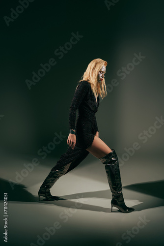 full length of stylish blonde woman with glitter on face walking in shiny dress on black backdrop