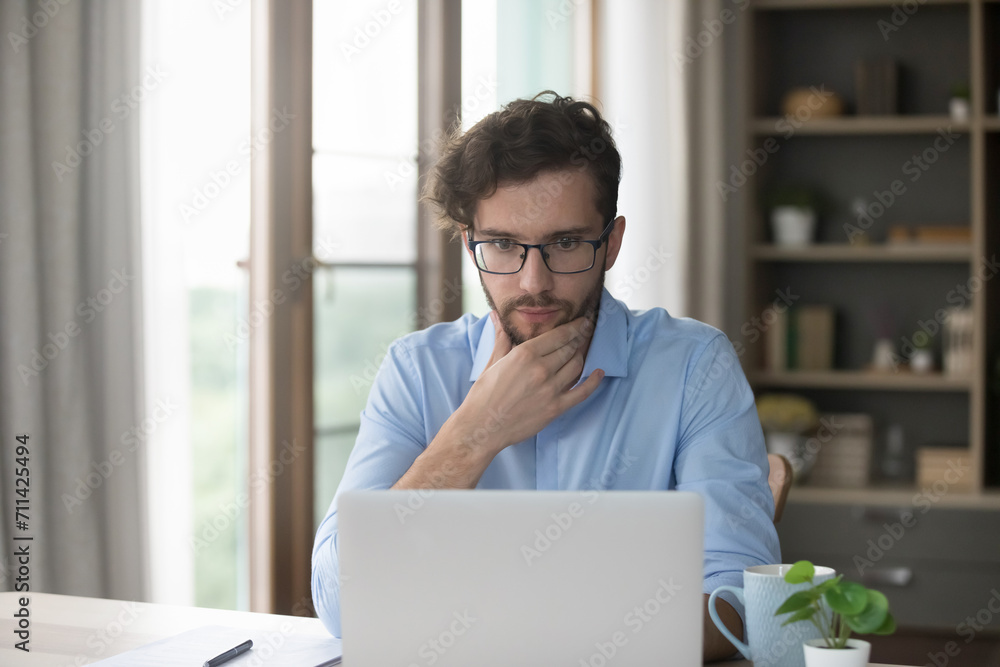 Serious thoughtful millennial man learn new artificial intelligence program, sit at desk staring at laptop, read received message, review information, consider business opportunity using modern device