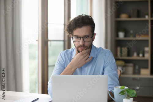 Serious thoughtful millennial man learn new artificial intelligence program, sit at desk staring at laptop, read received message, review information, consider business opportunity using modern device
