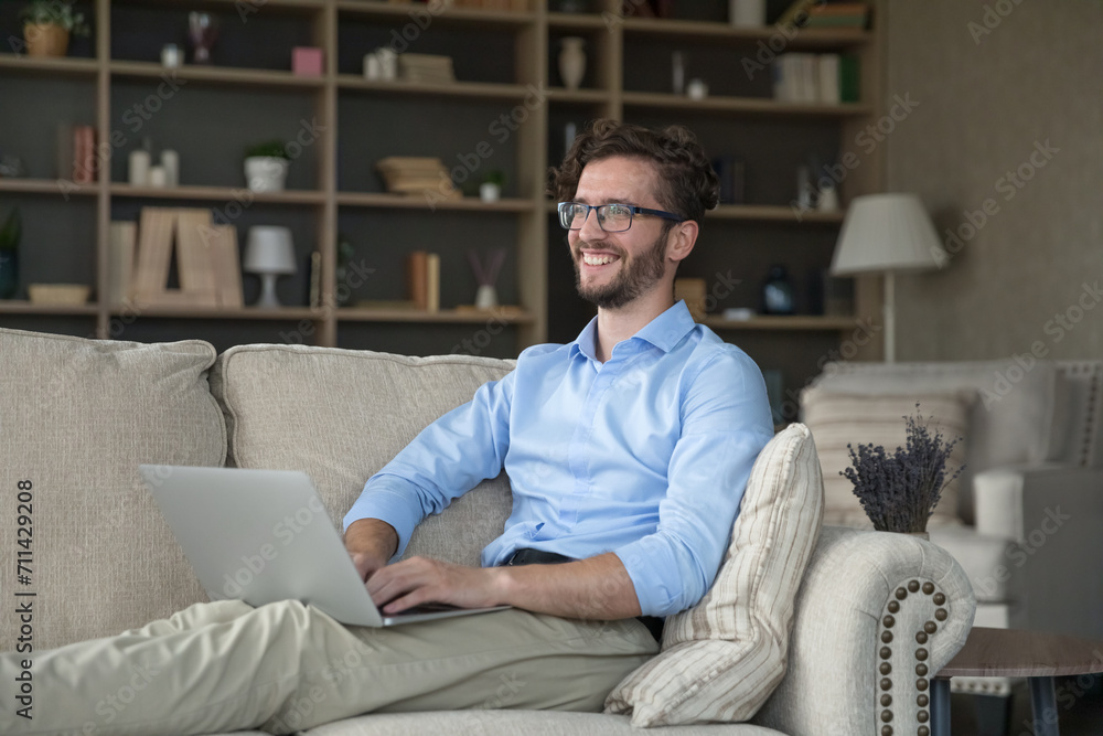 Happy young millennial man rest on sofa put wireless computer on laps smile, staring away, enjoy easy and comfortable e-shopping, share messages, solve business remotely spend leisure on internet Happy young millennial man rest on sofa put wireless computer on laps smile, staring away, enjoy easy and comfortable e-shopping, share messages, solve business remotely spend leisure on internet