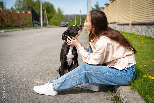 Fotografija Heartwarming image of a young girl sitting on the curb, sharing a tender moment as she kisses her affectionate pitbull breed dog